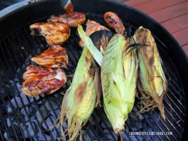 Three pieces of grilled unshucked corn, cooking with BBQ chicken on grill.