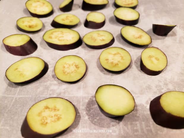 Baby eggplant slices are placed on a parchment paper-lined baking sheet.
