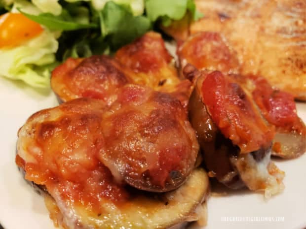 Some baby eggplant mozzarella bites on a white plate with salad behind them.