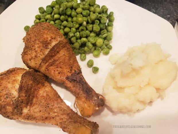 Two baked chicken legs on a plate with mashed potatoes and peas.