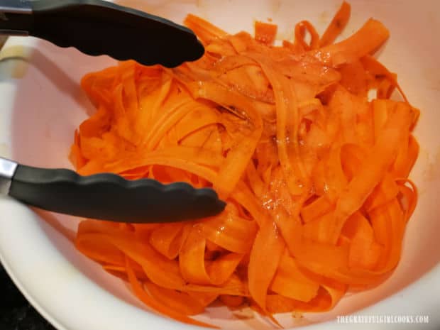 Tongs are used to combine carrot ribbons and salad dressing.