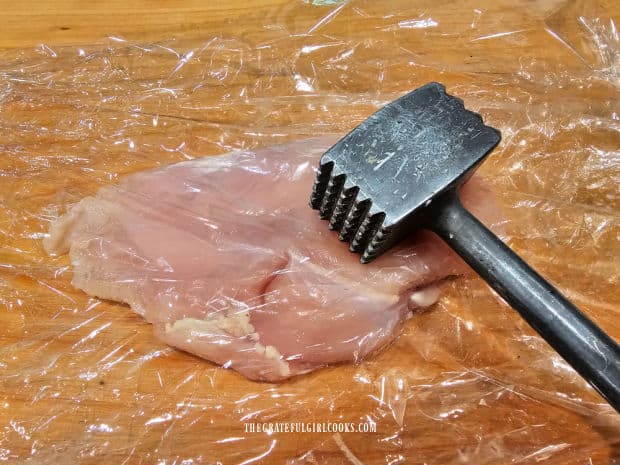 A chicken breast in plastic wrap is pounded flat with a meat mallet.