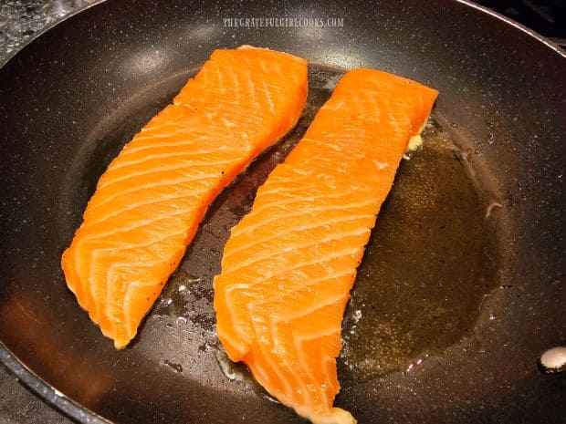 Two salmon fillets cook, seasoned side down, in hot oil in a skillet.