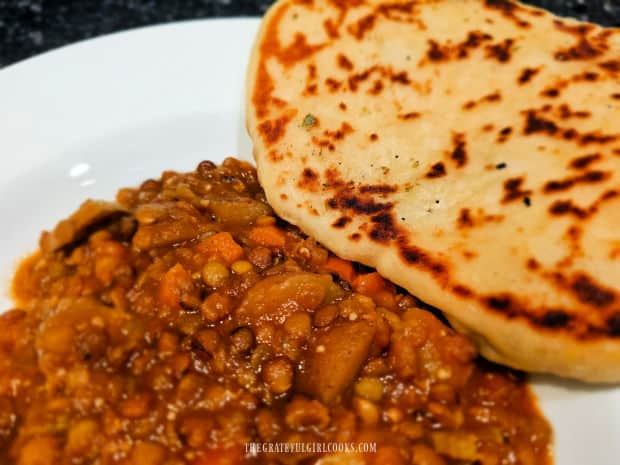 A white bowl of lentil potato soup, with garlic butter naan bread on the side.