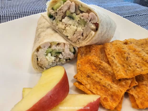 A white plate with two chicken wrap halves, apple slices and cheddar chips on the side.