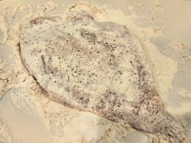 A chicken breast, coated in flour, seasoned with salt and pepper rests on a plate.