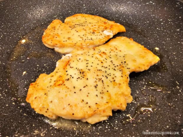 Two pan-seared chicken breasts cooking in olive oil in a skillet.