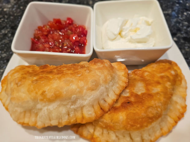 Two ground beef empanadas on a white plate, with sour cream and salsa for dipping.